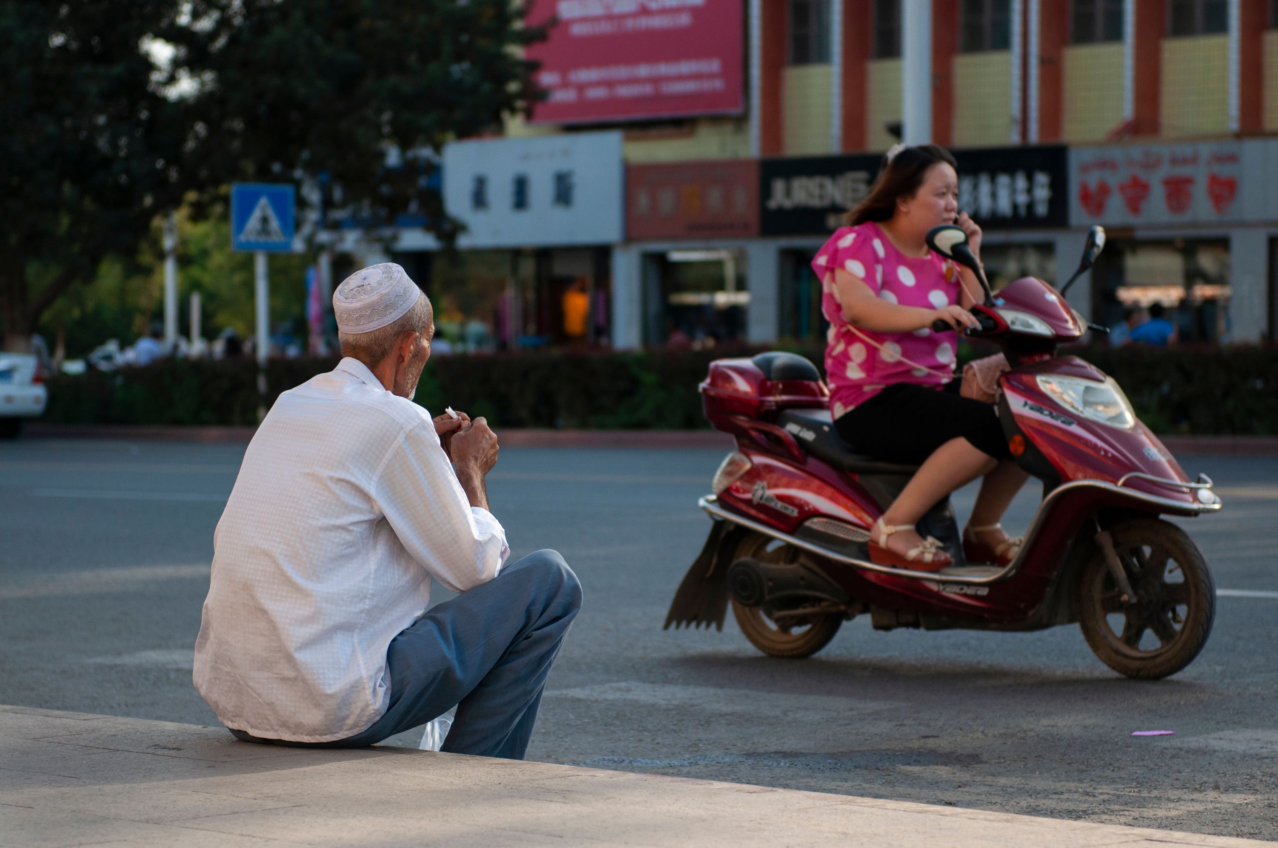 A Han Chinese woman riding a scooter and a Uyghur man sitting in a sidewalk
